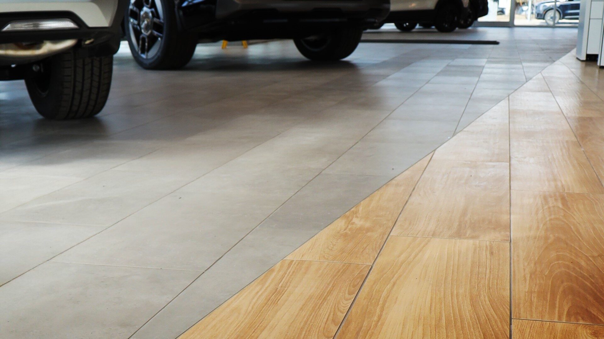 Flooring transition between gray tile and wood-look planks in a modern car dealership showroom.
